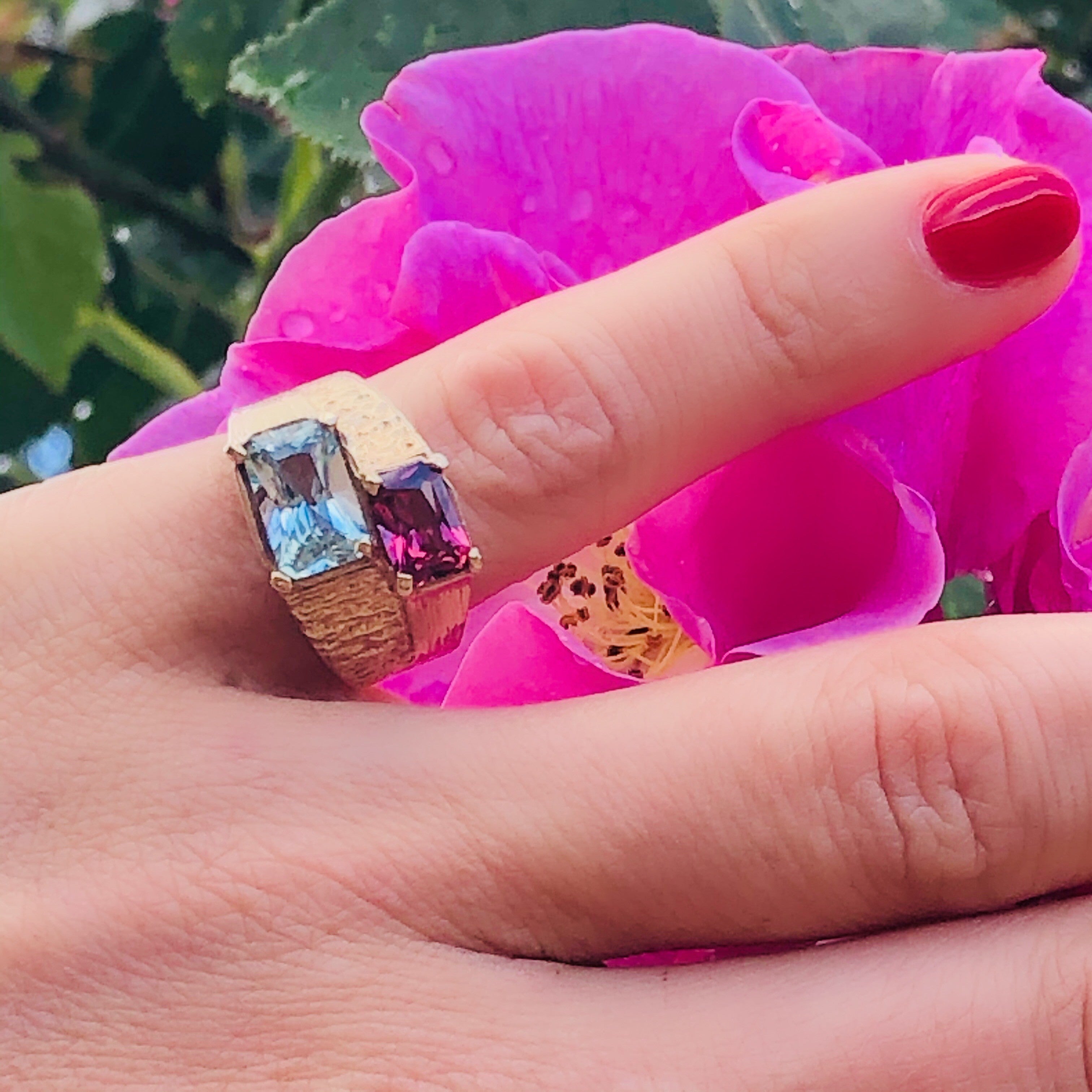 1970s Aquamarine and Pink Garnet Ring worn on models little finger. Models nail is painted red. Pink flower and green leaves in the background.