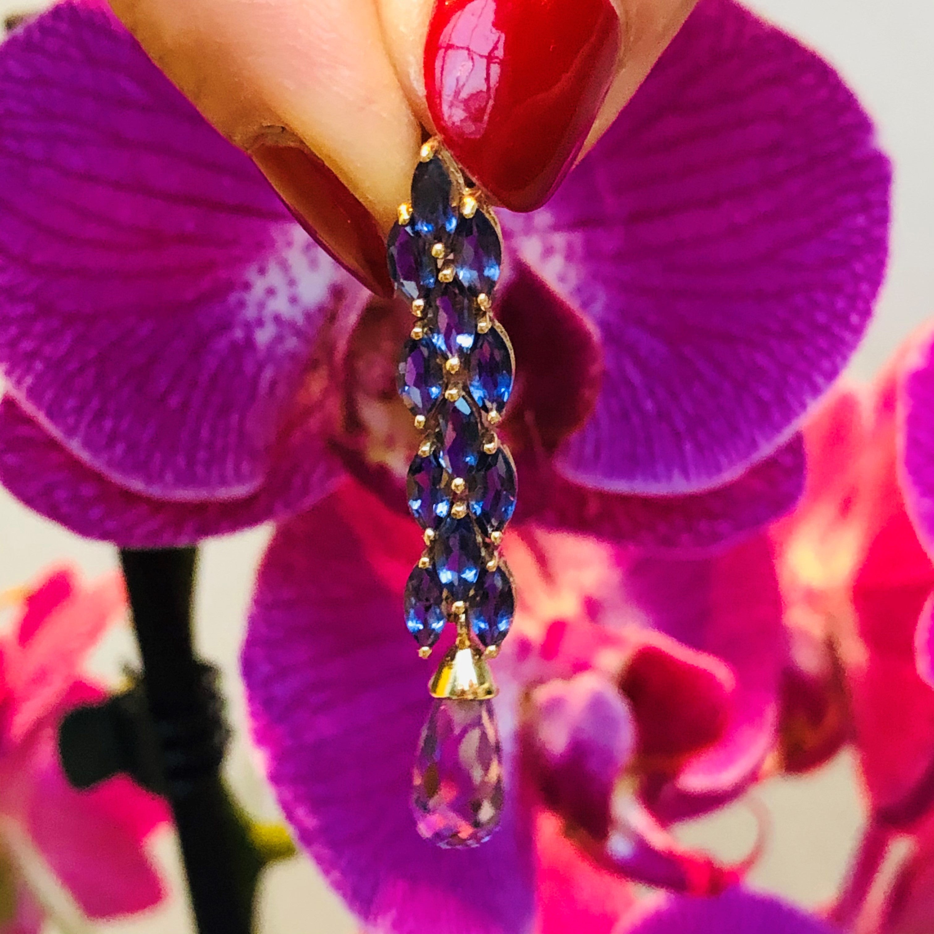 Alluring Amethyst & Iolite Cocktail Earring being held by models thumb and forefinger, models nails are painted a red pink colour photographed with a purple orchid flower in the background